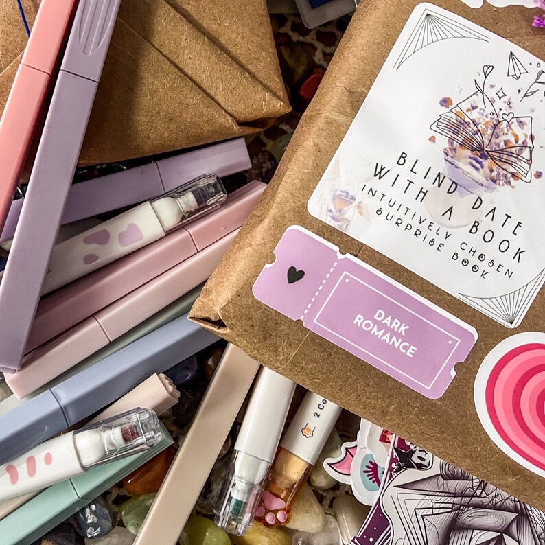 May include: A brown paper package with a sticker that says "Blind Date With A Book" and "Intuitively Chosen Surprise Book". The sticker is pink and has a heart on it. The package is surrounded by colorful markers and stickers.