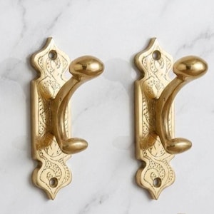 May include: Two brass wall hooks with an ornate, engraved design. Each hook has a curved arm and a rounded knob. The hooks are mounted on a decorative backplate with a floral pattern. The hooks are a warm gold color.