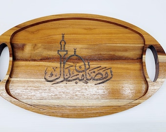 May include: Oval wooden serving tray with integrated handles. The tray features a laser-etched design of a mosque and Arabic calligraphy. The wood has a warm, natural tone with visible grain. A decorative piece for serving food or displaying items.
