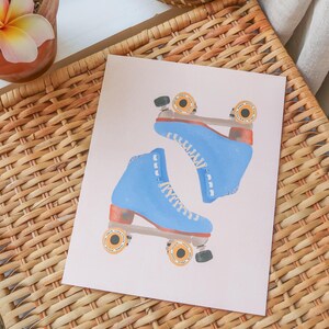 May include: A print featuring two blue roller skates with white laces and orange wheels. The skates are set against a light pink background, with a woven basket in the background.