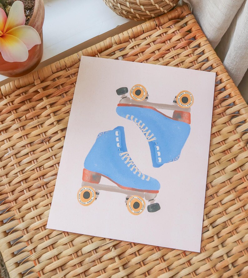 May include: A print featuring two blue roller skates with white laces and orange wheels. The skates are set against a light pink background, with a woven basket in the background.
