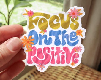 May include: A sticker with the words "Focus On The Positive" in yellow, blue, and pink, decorated with floral accents. The sticker is held by a hand, with a blurred background of greenery.