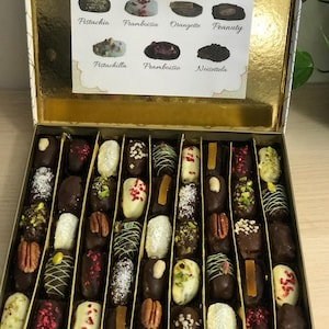 Könnte beinhalten: Eine goldene Schachtel gefüllt mit verschiedenen Pralinen, jede mit einzigartigen Belägen und Überzügen. Die Pralinen sind in Reihen angeordnet. Eine Karte zeigt die Geschmacksrichtungen: Amandine, Pecanila, Choco-coco, Pistachio, Framboisia, Orangette, Peanut, Pistachilla, Framboisia und Noisetella.