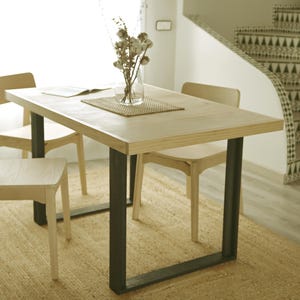 Peut inclure: Une table de salle à manger en bois clair avec des pieds en métal noir, entourée de quatre chaises en bois clair. Un vase en verre transparent avec des fleurs séchées est posé sur la table, avec un set de table tissé et un livre ouvert.