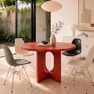 Puede incluir: Un comedor moderno con una mesa redonda roja y cuatro sillas de diferentes colores. Una lámpara colgante blanca cuelga encima. La habitación tiene grandes ventanales y una estética minimalista. La mesa mide aproximadamente 120 cm de diámetro.