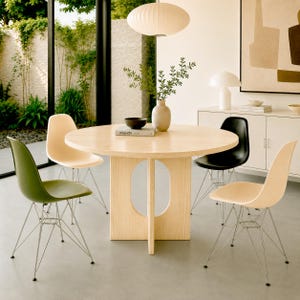 Puede incluir: Una mesa de comedor redonda de madera clara con una base central de corte ovalado, rodeada por cuatro sillas moldeadas en colores verde oliva, negro y crema, cada una con una base de alambre de metal fino. Un jarrón de cerámica con plantas.