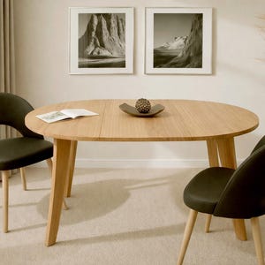 Op de afbeelding: Een ovale eikenhouten eettafel met twee zwarte stoelen. De tafel heeft een decoratieve schaal en een open boek. Twee ingelijste zwart-wit landschapsprints hangen aan de muur.