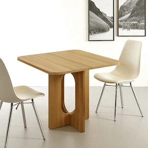Può includere: Tavolo quadrato in legno chiaro con angoli arrotondati e un supporto centrale unico con un ritaglio ovale. Due sedie color crema con gambe cromate lo affiancano. Il tavolo misura circa 76 cm per lato.