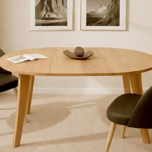 Op de afbeelding: Een ovale eettafel van licht hout met vier schuine poten. Twee zwarte stoelen met houten poten staan aan weerszijden van de tafel. Een decoratieve schaal en een open boek liggen op de tafel. Twee ingelijste landschapsprints hangen aan de muur.
