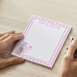 Może przedstawiać: Różowy notes z biało-różową kratką na obrzeżach. Notes ma różowy falisty brzeg i różowo-białe paski. Notes ma linię do pisania i tekst "fueled by Diet Coke" u góry. Na dole notesu znajduje się rysunek puszki Diet Coke z kwiatami.