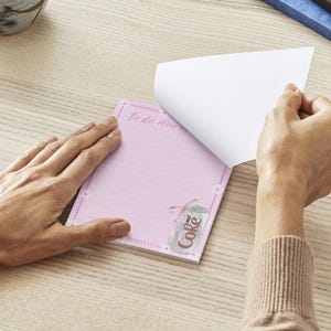 Peut inclure: Bloc-notes rose avec une liste de choses &agrave; faire et un graphique de canette de Coca-Cola. Le bloc-notes a des lignes roses et une bordure blanche.