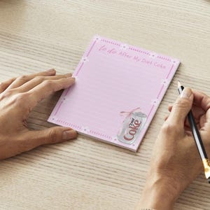 Peut inclure: Bloc-notes rose avec un int&eacute;rieur lign&eacute; et une bordure blanche. Le bloc-notes porte l'inscription "to do After My Diet Coke" en haut et un dessin d'une canette de Coca-Cola Light avec un ruban rose en bas.