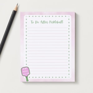 Op de afbeelding: Roze notitieblok met een aquarel achtergrond en een groene rand van hartjes. Het notitieblok heeft gestreept papier en de tekst "To Do After Pickleball" bovenaan. Een roze pickleball racket met de tekst "In My Pickleball Era" bevindt zich linksonder.