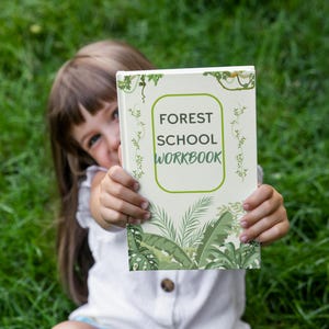May include: A young person is holding up a white workbook with green and brown illustrations. The cover reads "Forest School Workbook".