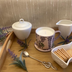 Könnte beinhalten: Ein Holztablett mit einer weißen Zuckerdose, einem weißen Milchkännchen, einer Kerze mit Blumenmuster in einer Tasse, einer kleinen Schale mit Keksen und Lavendelzweigen.