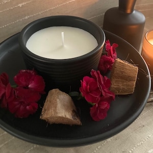 Puede incluir: Un portavelas de cerámica negro con una vela blanca y una mecha. El portavelas está sobre un plato negro con flores rojas y trozos de cáscara de coco.