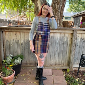 May include: A woman wearing a gray long-sleeved shirt and a plaid overall dress with a brown, blue, and red pattern. She is wearing black knee-high boots and has a tattoo on her leg that says "2002".