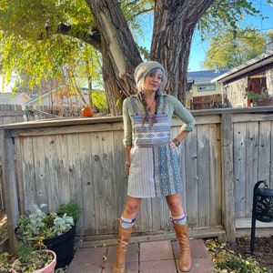 May include: A woman wearing a gray knit hat, a green shirt, a brown and white patterned overall dress, brown boots, and patterned socks. She is standing in front of a wooden fence with a tree behind her.