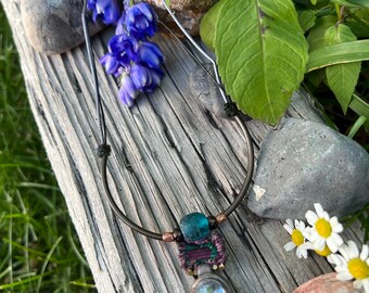 May include: A handmade necklace with a turquoise and purple pendant featuring a woven fabric design and a clear glass stone. The necklace is made with brown leather cord and a copper wire hoop.
