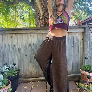 May include: A woman wearing a brown and black patterned wide-leg pants with a burgundy and multi-colored patchwork crop top. She is standing in front of a wooden fence.