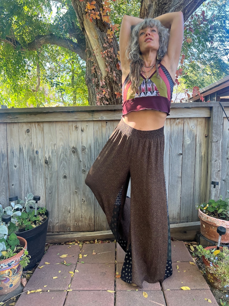 May include: A woman wearing a brown and black patterned wide-leg pants with a burgundy and multi-colored patchwork crop top. She is standing in front of a wooden fence.