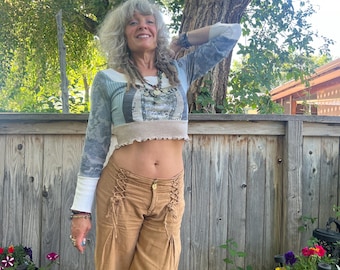May include: A cropped long-sleeve top with a patchwork design in shades of blue, white, and gray, paired with tan pants featuring lace-up detailing. The pants have a wide-leg cut. The outfit is worn outdoors.