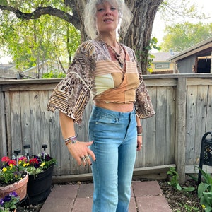May include: A woman wearing a brown and white patterned wrap top with bell sleeves and blue jeans. She is standing in front of a wooden fence with green plants in the background.