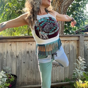 May include: A woman wearing a white halter top with a blue and brown graphic print of a motorcycle and the text "Crazy 8's" and "Black Hills Rally". The top has a fringe trim. She is also wearing blue and white patchwork pants with a checkered pattern at the bottom.