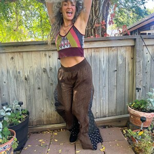 May include: A woman with gray hair is wearing a brown and black patterned pants with a multi-colored crop top. She is standing in front of a wooden fence with her arms raised in the air.