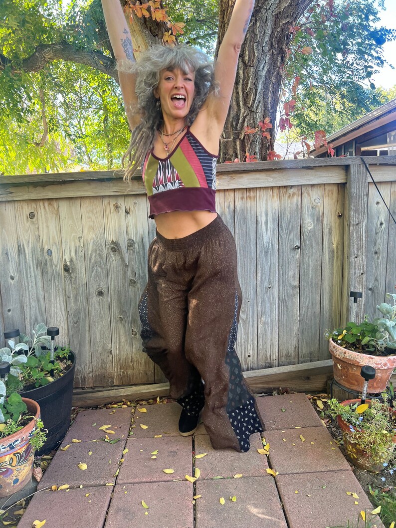 May include: A woman with gray hair is wearing a brown and black patterned pants with a multi-colored crop top. She is standing in front of a wooden fence with her arms raised in the air.