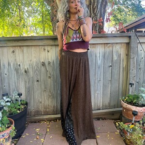 May include: A woman wearing a brown and black patterned pants with a maroon and green patterned crop top. She is standing in front of a wooden fence.