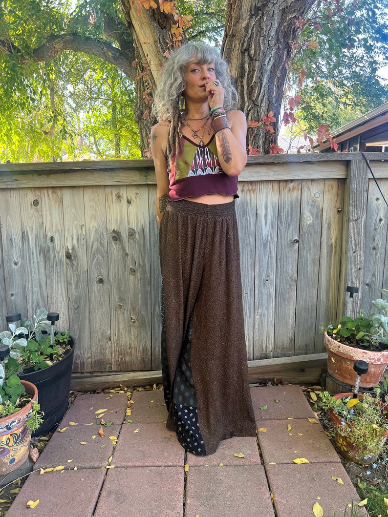 May include: A woman wearing a brown and black patterned pants with a maroon and green patterned crop top. She is standing in front of a wooden fence.