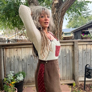 May include: A brown suede mini skirt with a red and white checkered pattern on the side. The skirt has a button closure and a small pocket on the front.