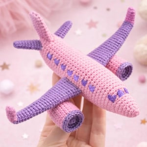 May include: A handmade crochet airplane in pink and purple. The body and wings are pink, with purple accents on the wings and tail. Small purple hearts decorate the body. A soft, whimsical toy.