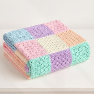 Puede incluir: Una manta de ganchillo plegada, de colores pastel. La manta está compuesta por cuadrados en tonos azul claro, rosa, melocotón, verde menta, lavanda y crema. Los cuadrados presentan varios patrones de ganchillo texturizados. La manta está doblada cuidadosamente sobre una superficie blanca.