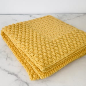 Può includere: Coperta gialla all'uncinetto piegata con un motivo a pois in rilievo. La coperta è realizzata in filato morbido ed è piegata ordinatamente, mostrando la dettagliata lavorazione a punto. Il colore è un giallo caldo e solare.