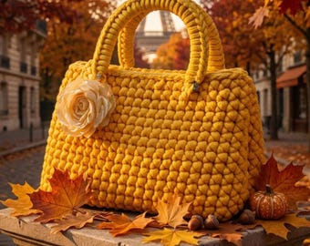 May include: A bright yellow crocheted handbag featuring a thick braided handle and a large, pale cream-coloured fabric rose attached to the front. The bag has a dense, textured stitch pattern and is displayed with autumn leaves and a small pumpkin.