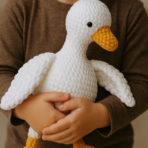 May include: A white crochet goose plush toy with a yellow beak and feet is held by a child. The handmade stuffed animal has black eyes and is made of soft, textured yarn. The goose has outstretched wings and is a cuddly toy.