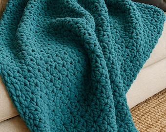 Op de afbeelding: Een teal-kleurige, dik gebreide deken gedrapeerd over een beige bank. De deken heeft een gestructureerd, gelust patroon, waardoor het er gezellig en uitnodigend uitziet. Een donkergrijs kussen ligt op de achtergrond. Ideaal voor interieurdecoratie.