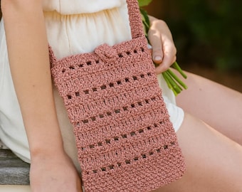 Puede incluir: Un bolso cruzado de ganchillo rosa empolvado con una correa y un cierre de botón. El bolso tiene forma rectangular con un diseño de tejido abierto y texturizado. La correa también está tejida a ganchillo. El bolso mide unos 20 cm de largo y 13 cm de ancho.