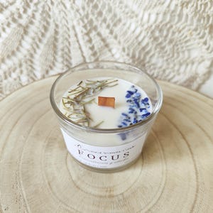 May include: A clear glass candle with a white wax. The candle is decorated with dried rosemary, blue flowers, and a wooden wick. The label reads "FOCUS". The candle sits on a wooden slice.
