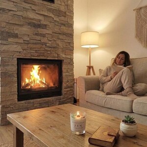Puede incluir: Una acogedora sala de estar con una chimenea encendida, una mesa de centro de madera y una mujer leyendo en un sofá beige. Una vela con la palabra "ZEN" está sobre la mesa, junto con una pequeña planta en maceta y un libro. Una lámpara proporciona una iluminación suave.