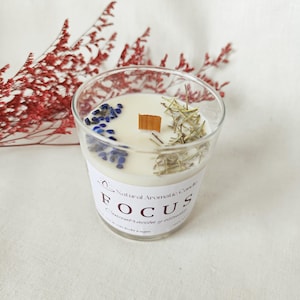 May include: A clear glass candle with a white wax interior, decorated with blue stones, dried herbs, and a wooden wick. The candle is labelled "FOCUS" with additional text, and is a natural aromatic candle.