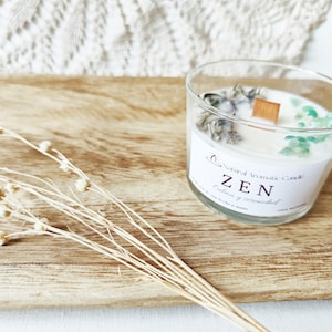 Op de afbeelding: Een heldere glazen kaars met het woord "ZEN" op het etiket, omringd door decoratieve stenen en een houten lont. De kaars staat op een houten dienblad met gedroogde bloemen.