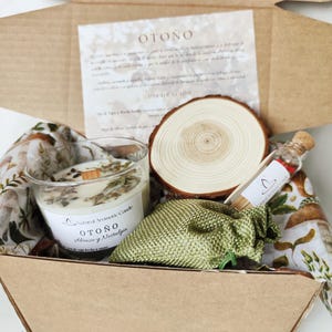 May include: A cardboard gift box containing a glass candle with a wooden wick and dried herbs, a round wooden slice, a small green fabric pouch, and a glass vial with a cork stopper. A printed card with the text OTOÑO is included inside.