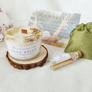 Può includere: Una candela di cera di soia in un bicchiere trasparente con uno stoppino di legno, fiori secchi e il testo "Buen Rollo". La candela è su un sottobicchiere di legno, con un piccolo sacchetto verde e una bottiglia di fiammiferi.
