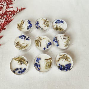 Può includere: Nove piccole candele a forma di lumino bianche in supporti argentati. Ogni candela è decorata con pietre blu ed erbe secche. Le candele sono disposte su una superficie bianca, con fiori secchi rossi sullo sfondo.