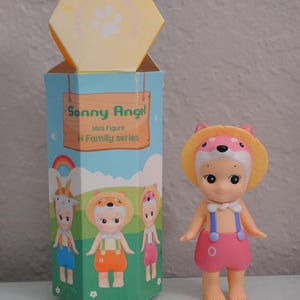 May include: A Sonny Angel mini figure from the H Family series, featuring a doll with a pink outfit and a yellow hat with a pink bear design. The box is light blue with illustrations of other figures.