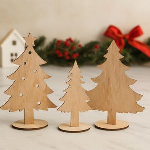 Peut inclure: Trois décorations de sapins de Noël en bois de différentes tailles. Les arbres sont découpés dans du bois de couleur claire et ont une finition naturelle. Le plus grand arbre a une étoile sur le dessus et des trous décoratifs. Chaque arbre repose sur une base ronde en bois.