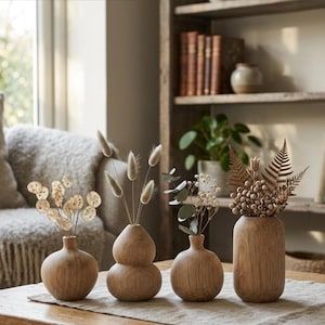 Könnte beinhalten: Vier Holzvasen in verschiedenen Formen, jede mit getrockneten Blumen und Blättern. Die Vasen sind auf einem neutralfarbenen Tischläufer angeordnet. Ein Bücherregal und ein Sofa sind im Hintergrund zu sehen.